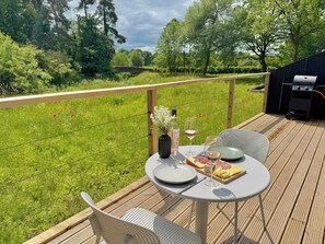 A wooden deck with a table set for a meal, featuring a bottle of wine, glasses, and a plate of food. - Luxurious Eco-Retreat in Market Drayton (Market Drayton)