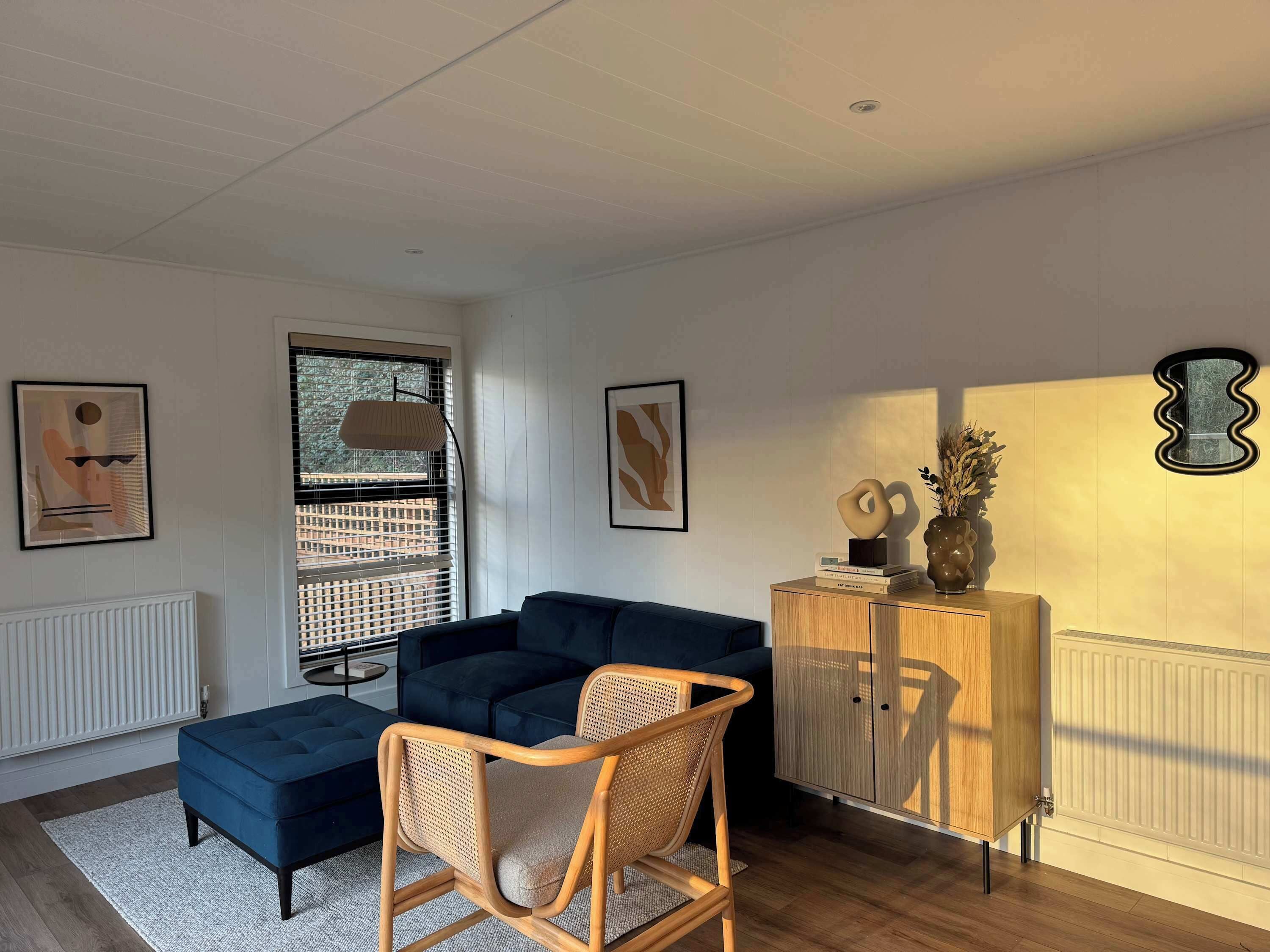A modern living room with a dark blue sectional sofa, a wicker chair, a wooden cabinet with decorative items, and abstract wall art.