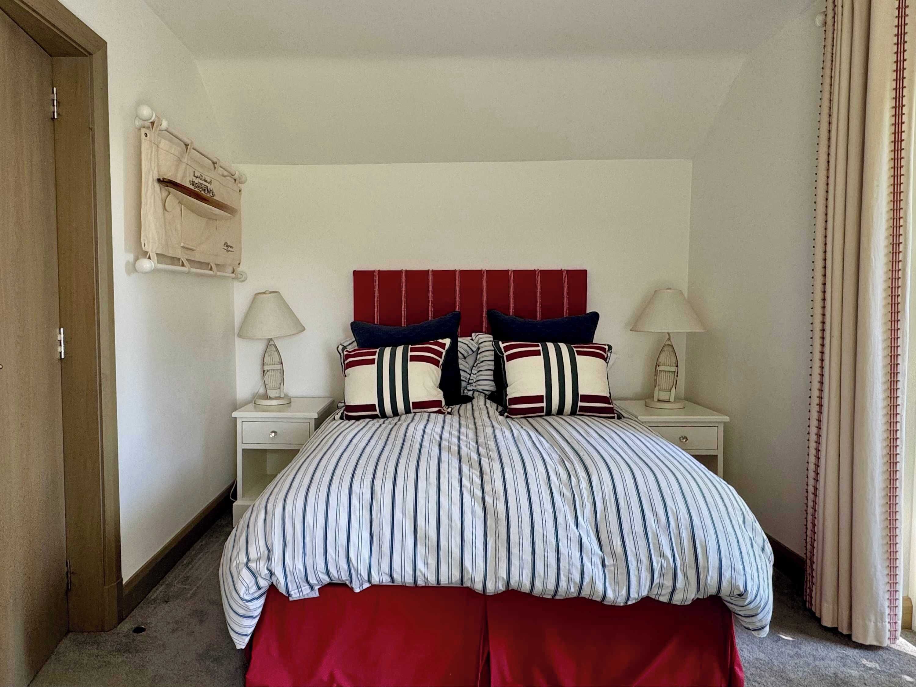 A bedroom with a striped bedspread, red headboard, and matching red curtains. There are two bedside tables with lamps and a small decorative item on each.