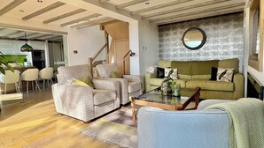 Ein modernes Wohnzimmer mit einer Couch, Sesseln und einem Couchtisch. Eine Treppe führt zu einer weiteren Ebene, und im Hintergrund ist eine Küchenzeile erkennbar.