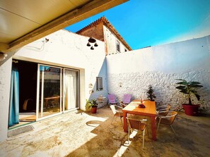 Une terrasse avec une table et des chaises en bois, des plantes en pot et une porte-fenêtre coulissante donnant sur un espace intérieur.