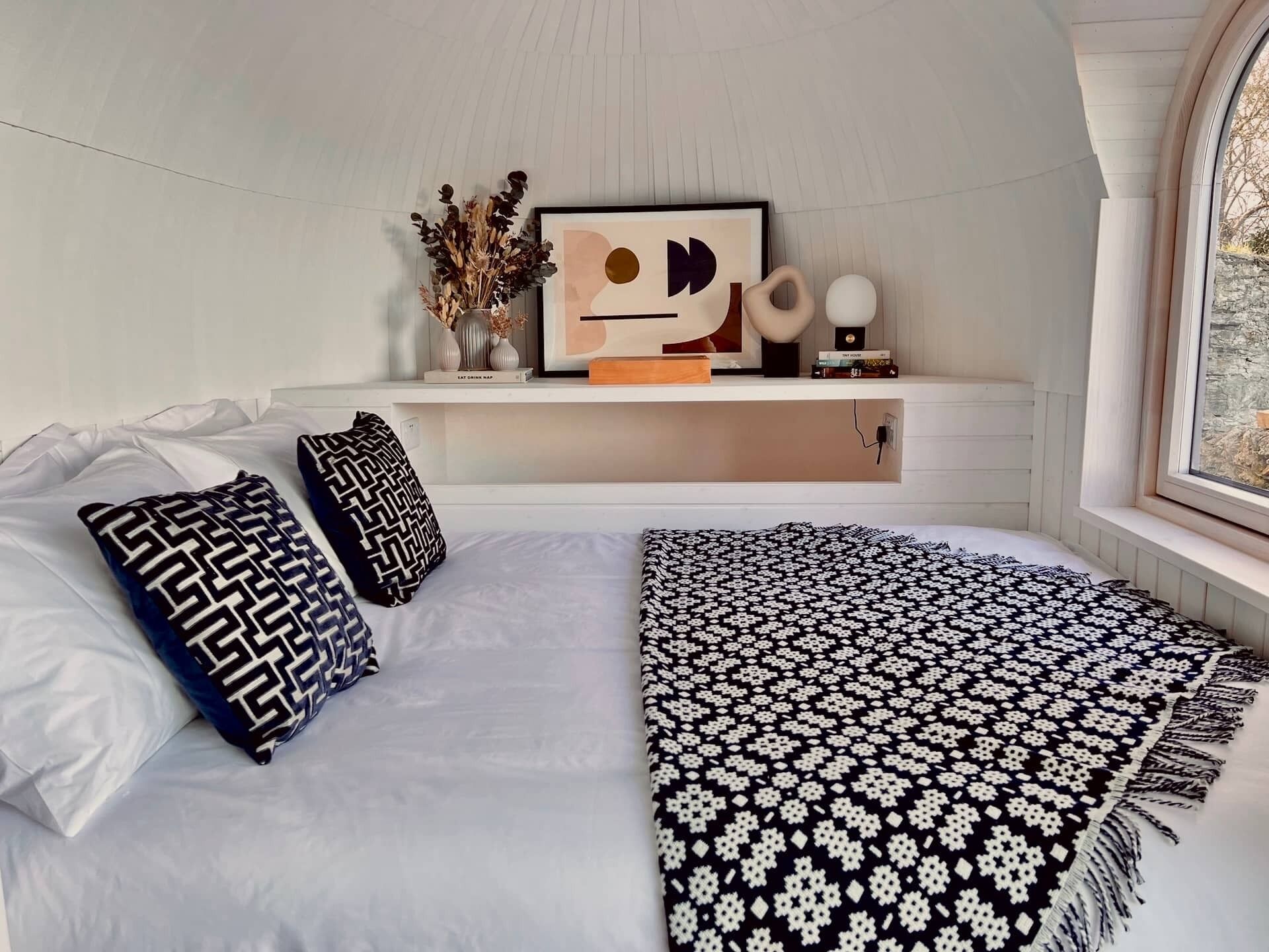 A bedroom with a bed, pillows, a bedside table with a vase, books, and a framed picture.