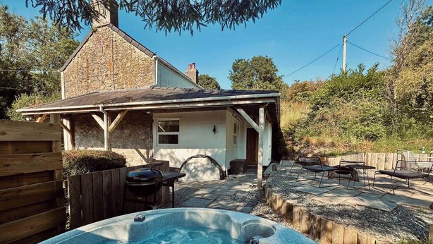 A hot tub in a backyard with a wooden deck, a covered patio, and a stone house.