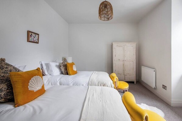 A bedroom with two beds, a white wardrobe, a yellow armchair, and a wicker pendant light.