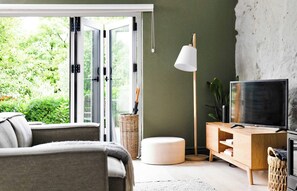 A living room with a sofa, a TV, a floor lamp, a wooden cabinet, and a large window with a view of greenery. - Nordic Stone Bothy with Sauna · Anglesey (Llangefni)