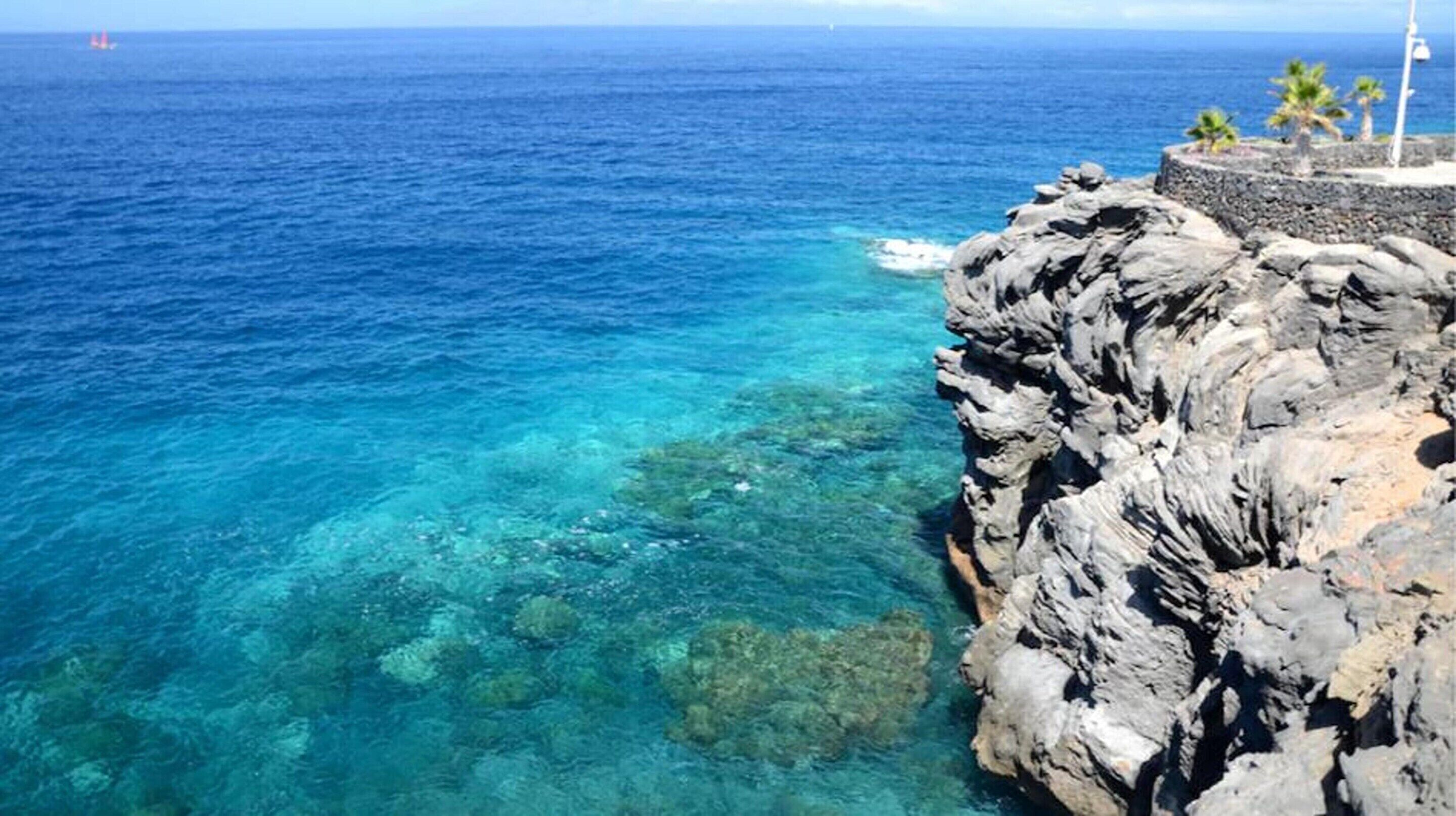 Une côte rocheuse bordée d’une eau d’un bleu limpide, avec un voilier qui se profile à l’horizon.