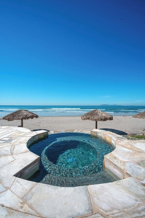 Un cielo azul despejado, una playa y una piscina con escalones de piedra y sombrillas de paja.