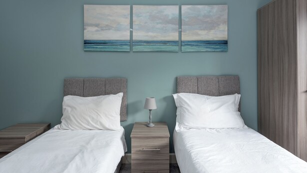A bedroom with two beds, a nightstand with a lamp, and a wall-mounted artwork of a seascape.
