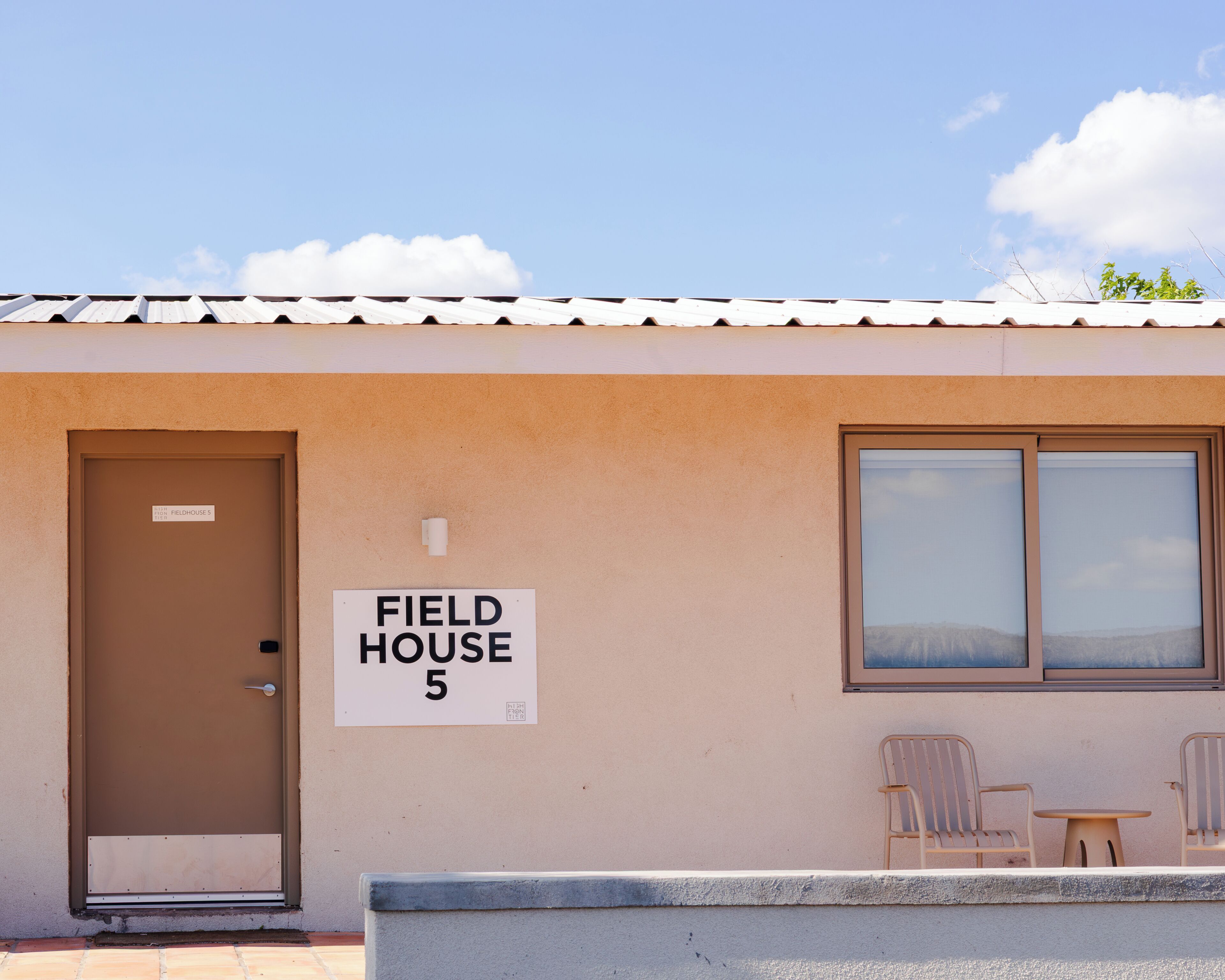 Un edifici amb un rètol que diu 'FIELD HOUSE 5' i una porta. Hi ha dues cadires i una taula petita a l'exterior.