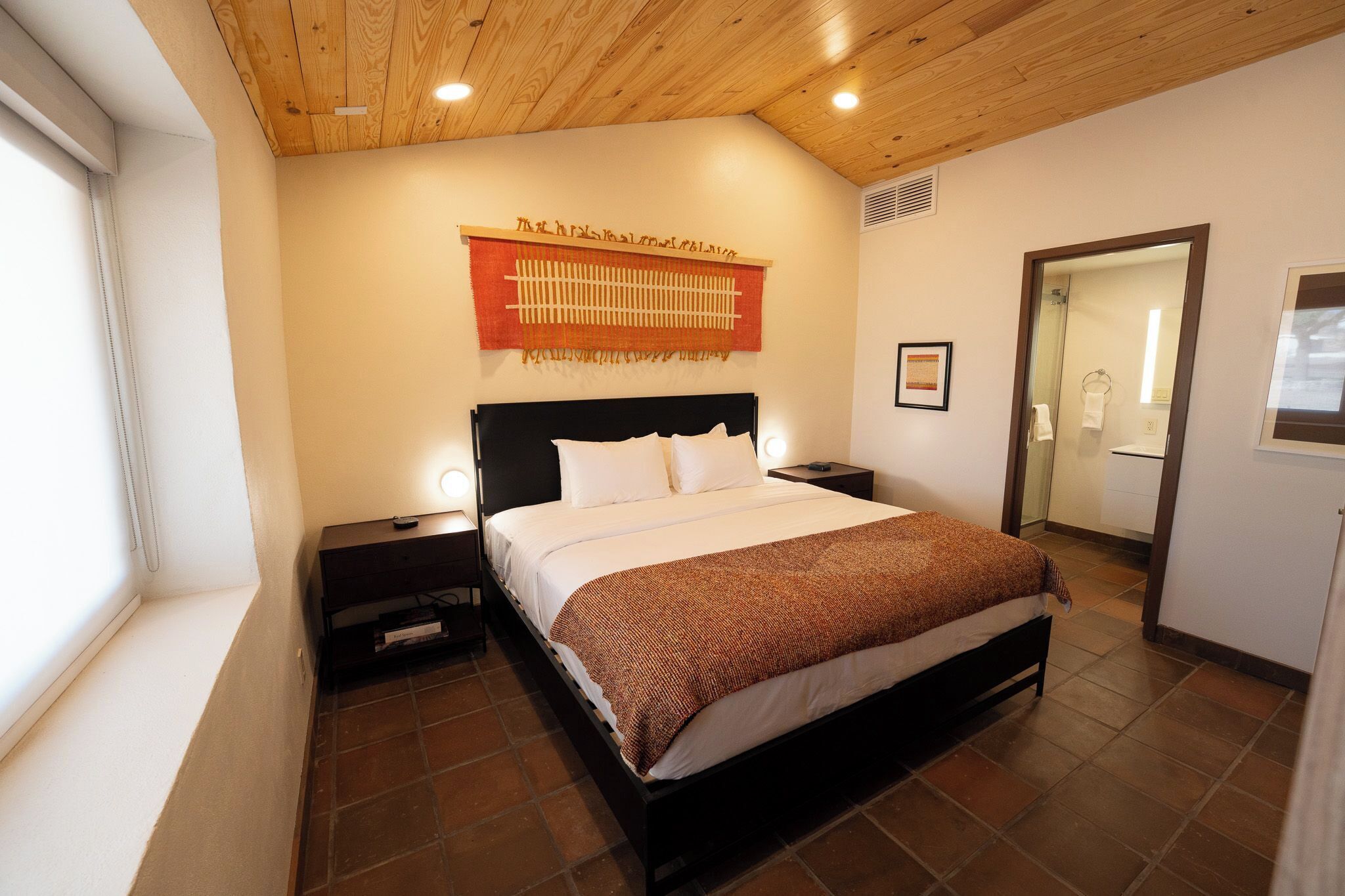 Um quarto com teto de madeira, uma cama com roupa de cama branca e manta marrom, uma mesa de cabeceira, uma obra de arte na parede e um banheiro visível através de uma porta aberta.