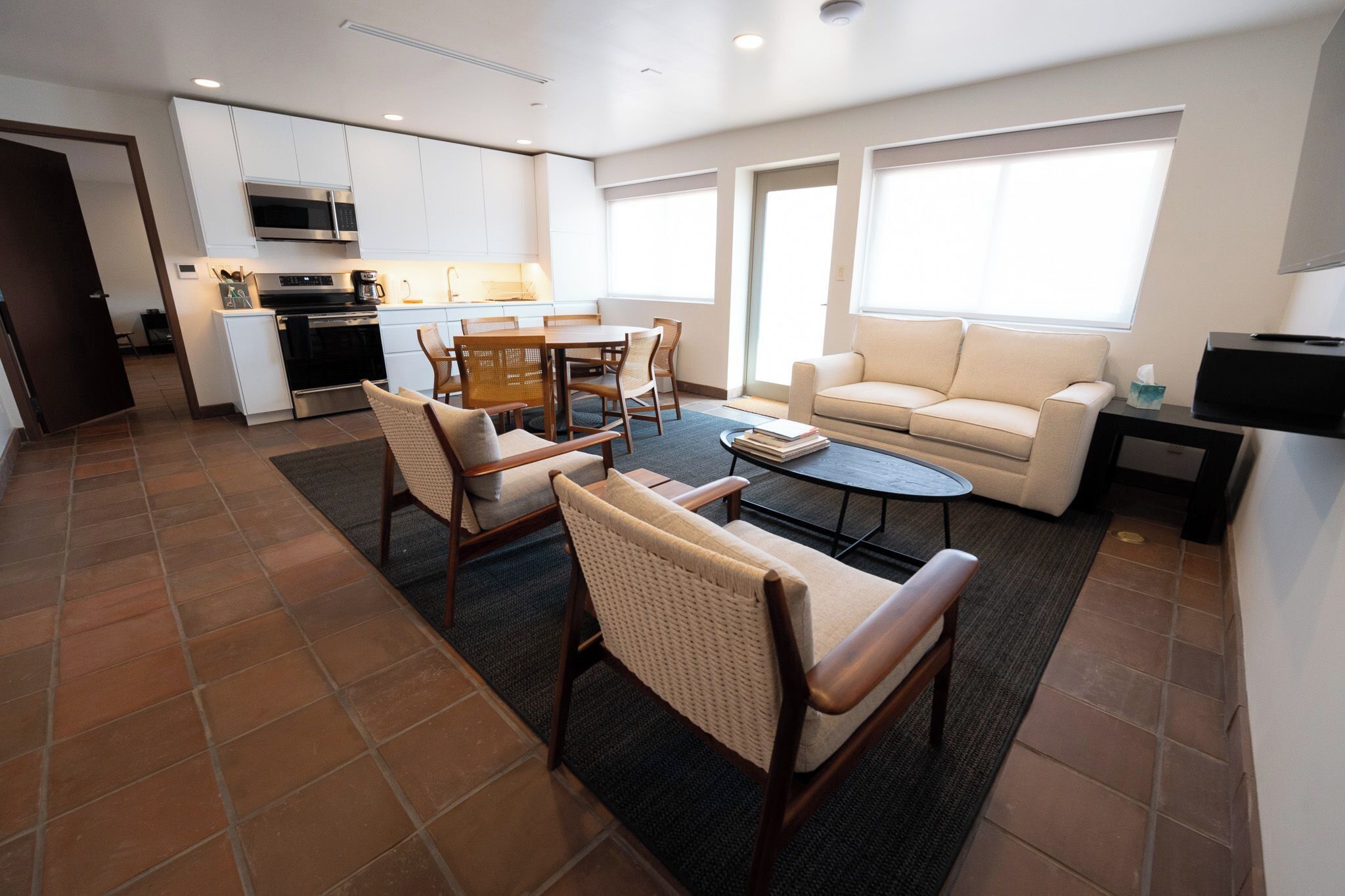 Un salon moderne comprenant un canapé, deux fauteuils, une table basse et un coin repas avec une table et des chaises. On aperçoit une cuisine équipée d’un four à micro-ondes et d’un four au fond de la pièce.