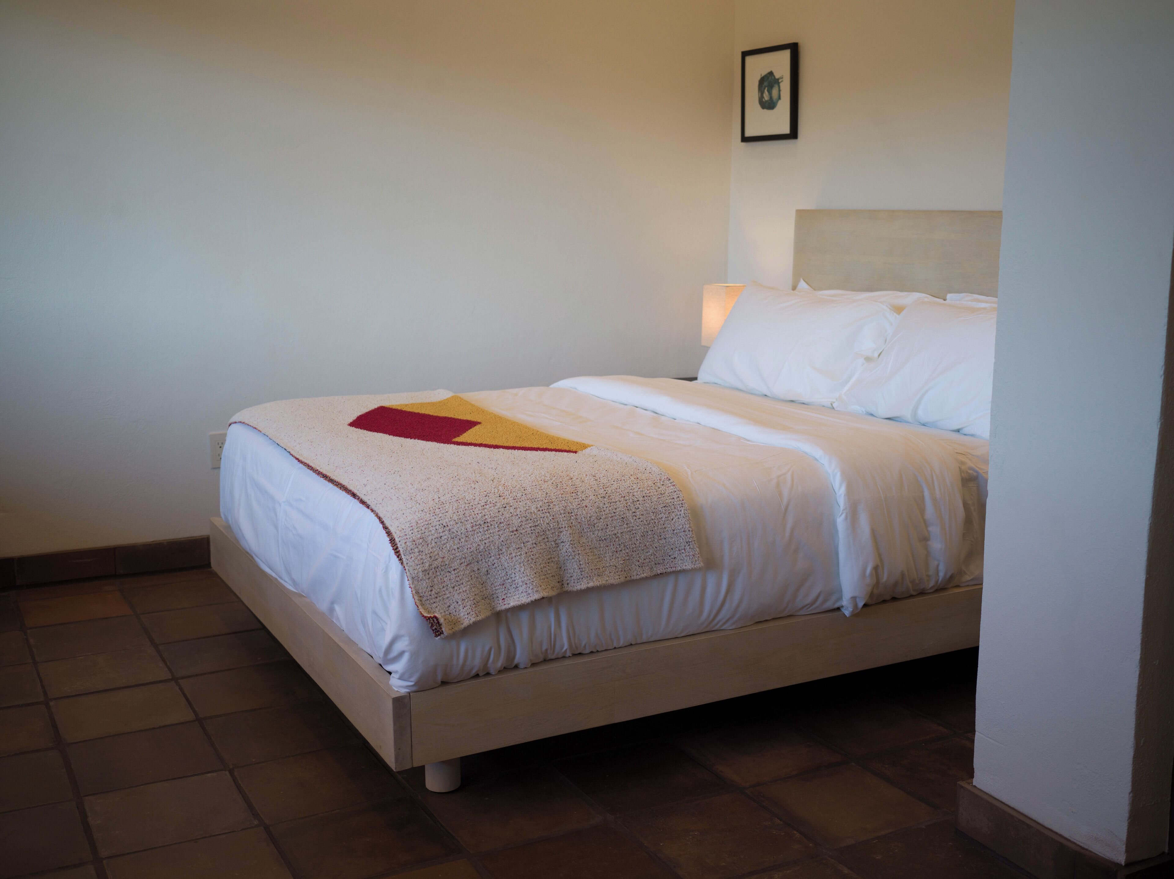 Un lit bien fait, avec une simple couverture et des oreillers, un petit tableau accroché au mur et un sol carrelé sobre.