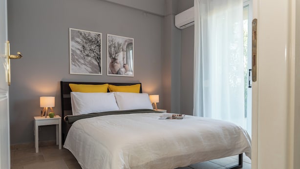 A neatly made bed with white and yellow bedding, two bedside tables with lamps, and two wall-mounted artworks depicting trees and architectural elements. - Stay & Smile apartments (Kallithea)