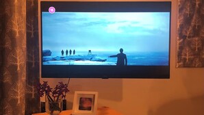 Ein an der Wand befestigter Fernseher, davor ein Holztisch, eine Vase mit Blumen und ein Bilderrahmen.