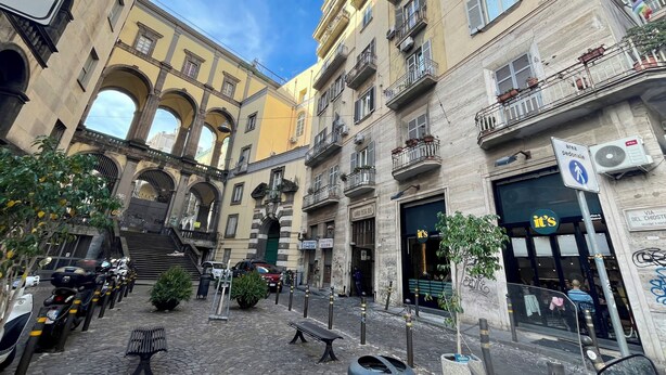 Un patio empedrado con edificios históricos, una señal que indica zona peatonal y una tienda con un letrero que dice 'it's'.