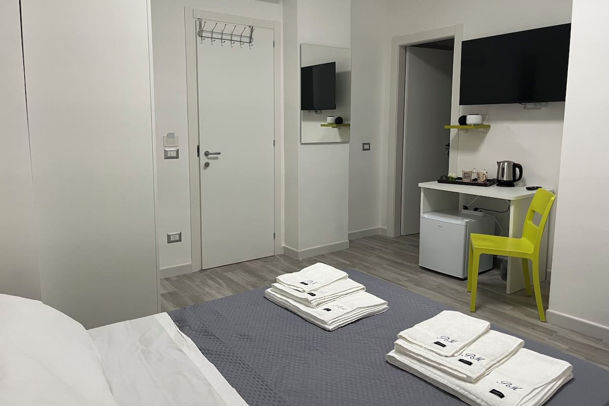 A hotel room with a bed, a desk with a chair, a TV, and a shelf with items.