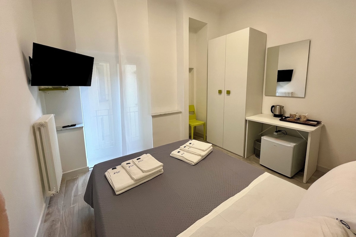 A hotel room with a bed, a desk with a chair, a mirror, a radiator, and a television mounted on the wall.