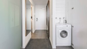 A narrow hallway with a washing machine and a mirror on the left, a door at the end, and a tiled floor.