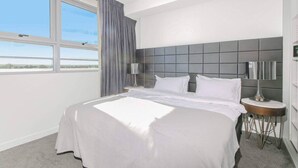 A hotel room with a large bed, a headboard with a grey panel, two bedside tables with lamps, a window with curtains, and a view of the outdoors.