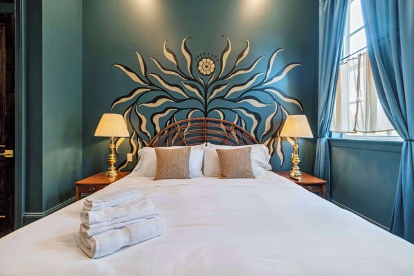 Una cama bien tendida con un cabecero decorativo, dos lámparas de noche y una ventana con cortinas.