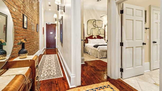 Un pasillo con pared de ladrillo, banca de madera y un tapete que conduce a un dormitorio con cama y cabecera.