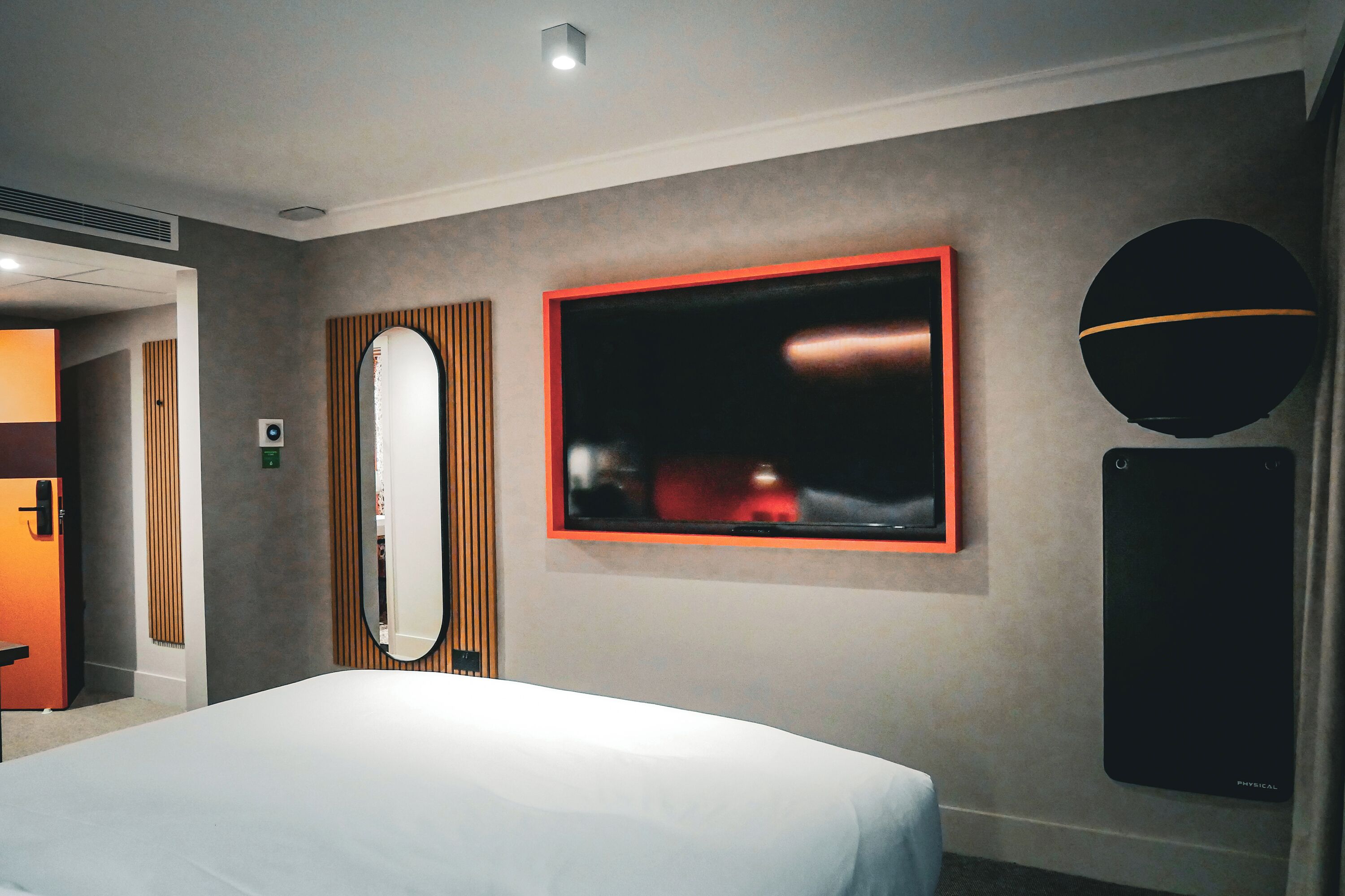 A hotel room with a bed, a large mirror, a flat-screen TV, and a wall-mounted speaker.
