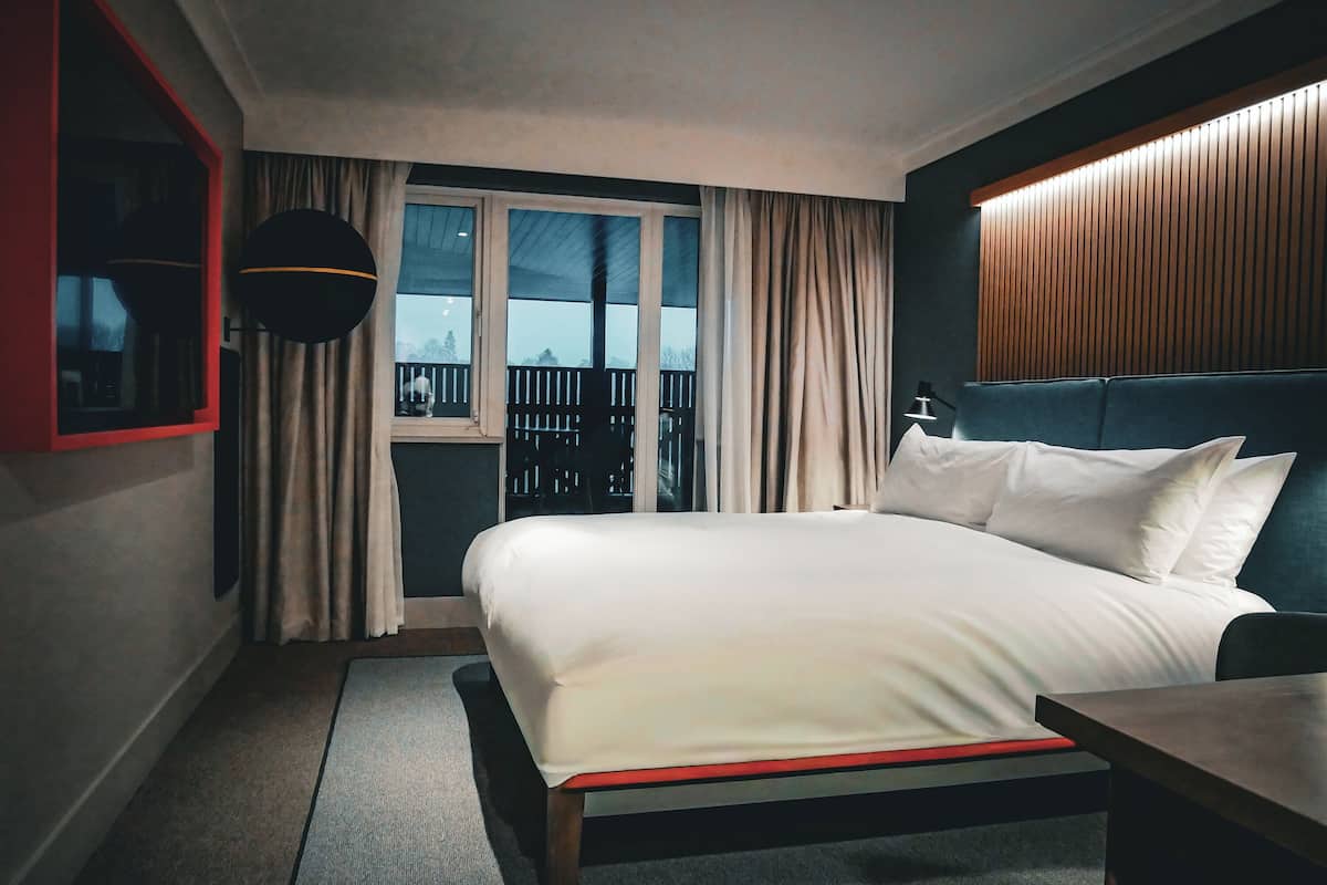 A hotel room with a large bed, a small table, a window with curtains, a balcony view, and a wooden headboard.