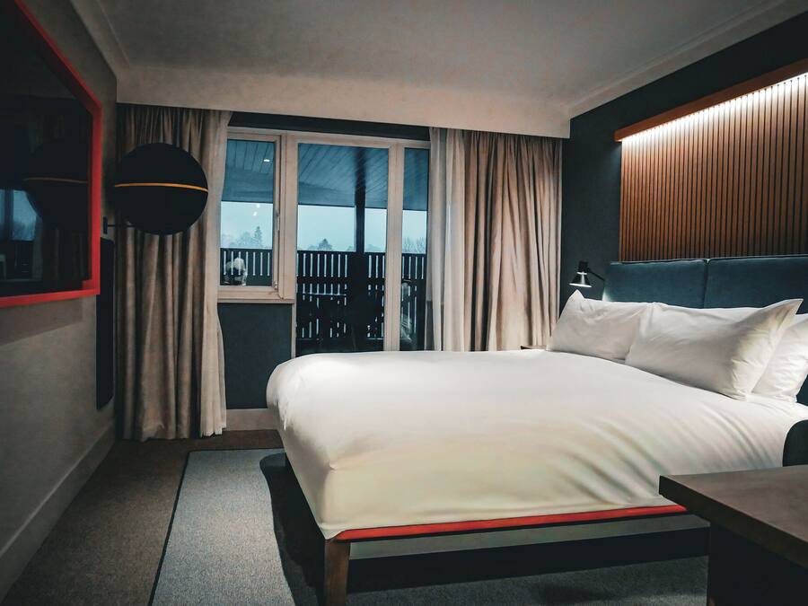 A hotel room with a large bed, a small table, a window with curtains, a balcony view, and a wooden headboard.
