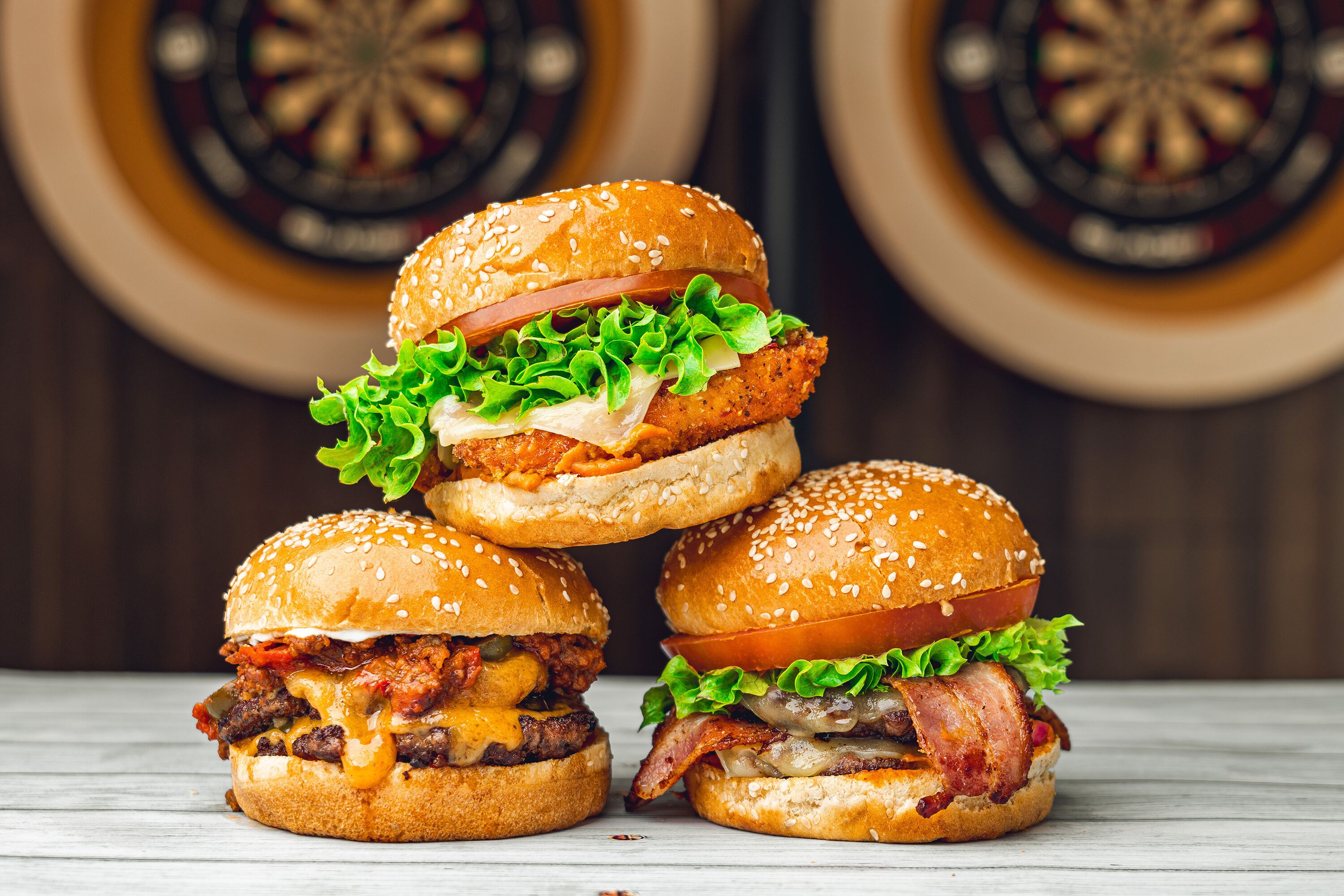 Tres hamburguesas apiladas sobre una superficie de madera, con dardos y un tablero de dardos al fondo.