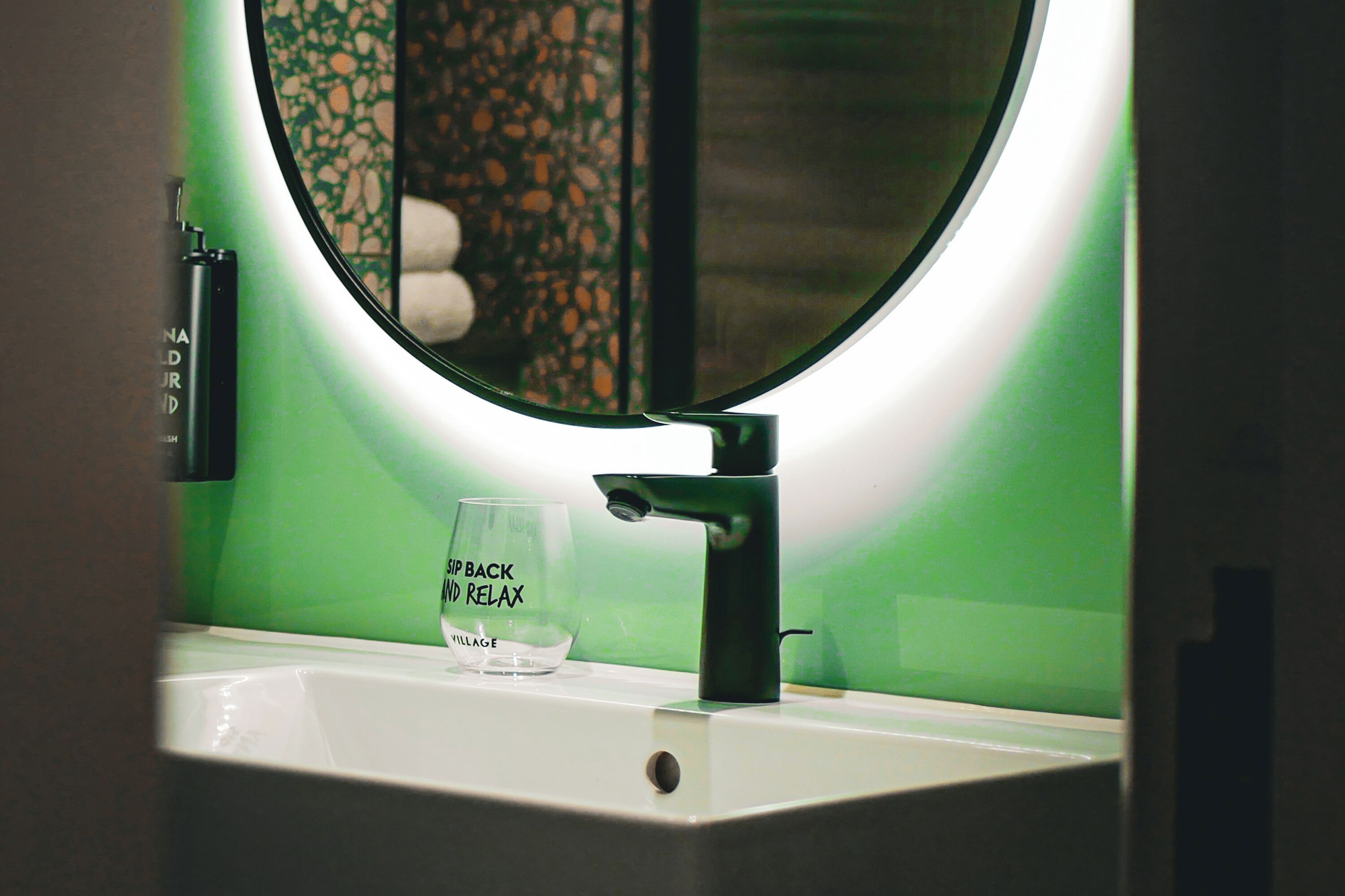 A bathroom sink with a glass reading "Sip Back and Relax" and a mirror with a circular light.