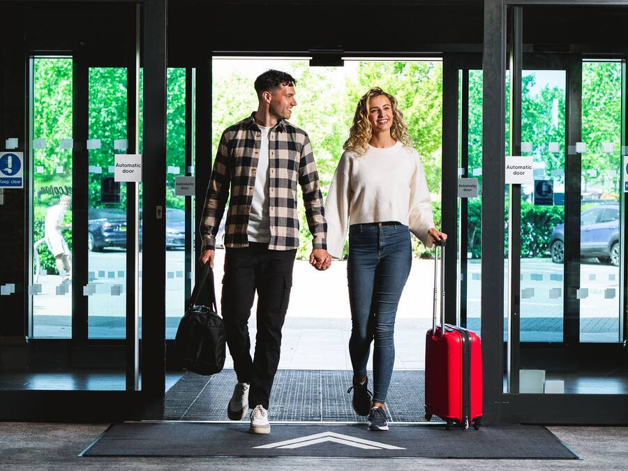 Una pareja caminando por una puerta automática con una maleta roja, un bolso negro y un felpudo en el suelo.
