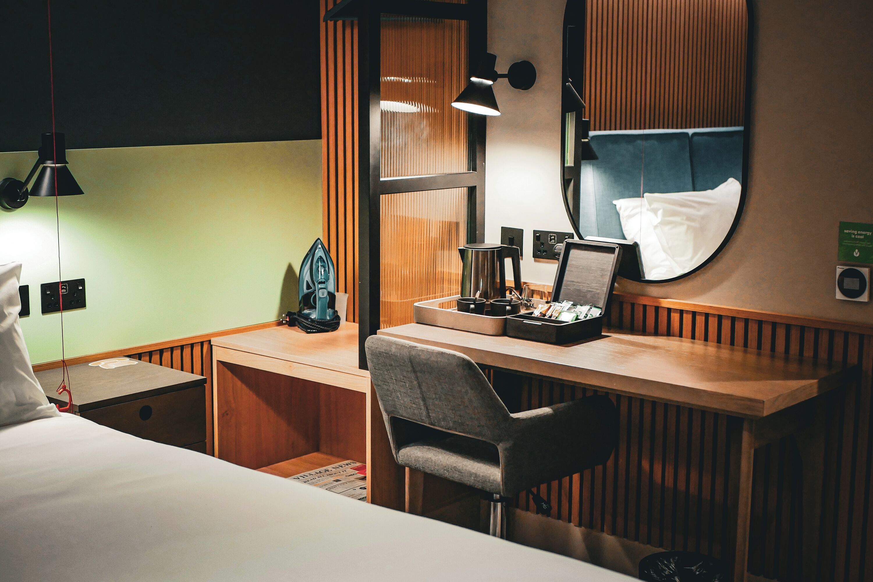 A hotel room with a bed, a desk with a chair, a mirror, and a small table with a kettle and cups.