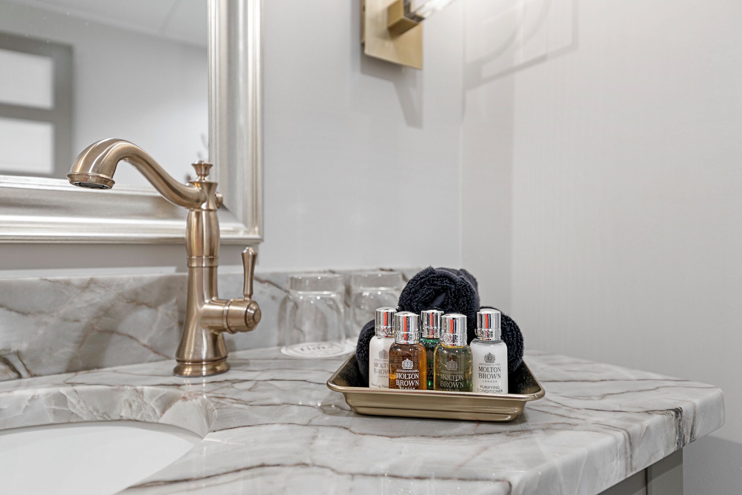 A bathroom with a marble countertop, a gold faucet, and a tray with toiletries including bottles and a towel.