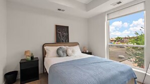 Une chambre avec une grande fenêtre, un lit recouvert d’une couverture bleue, une table de chevet avec une lampe et un tableau encadré accroché au mur.