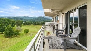 A balcony with two Adirondack chairs, a small table, and a view of a green landscape with trees and hills. - Penthouse Paradise | Smoky Mtn Views & Jacuzzi (Pigeon Forge)
