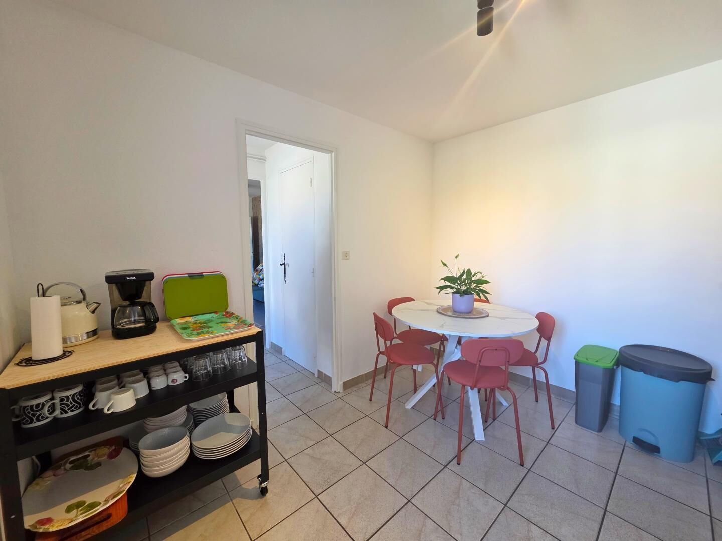 A small dining area with a round table and red chairs, a coffee maker, a kettle, and a shelf with dishes and cups.