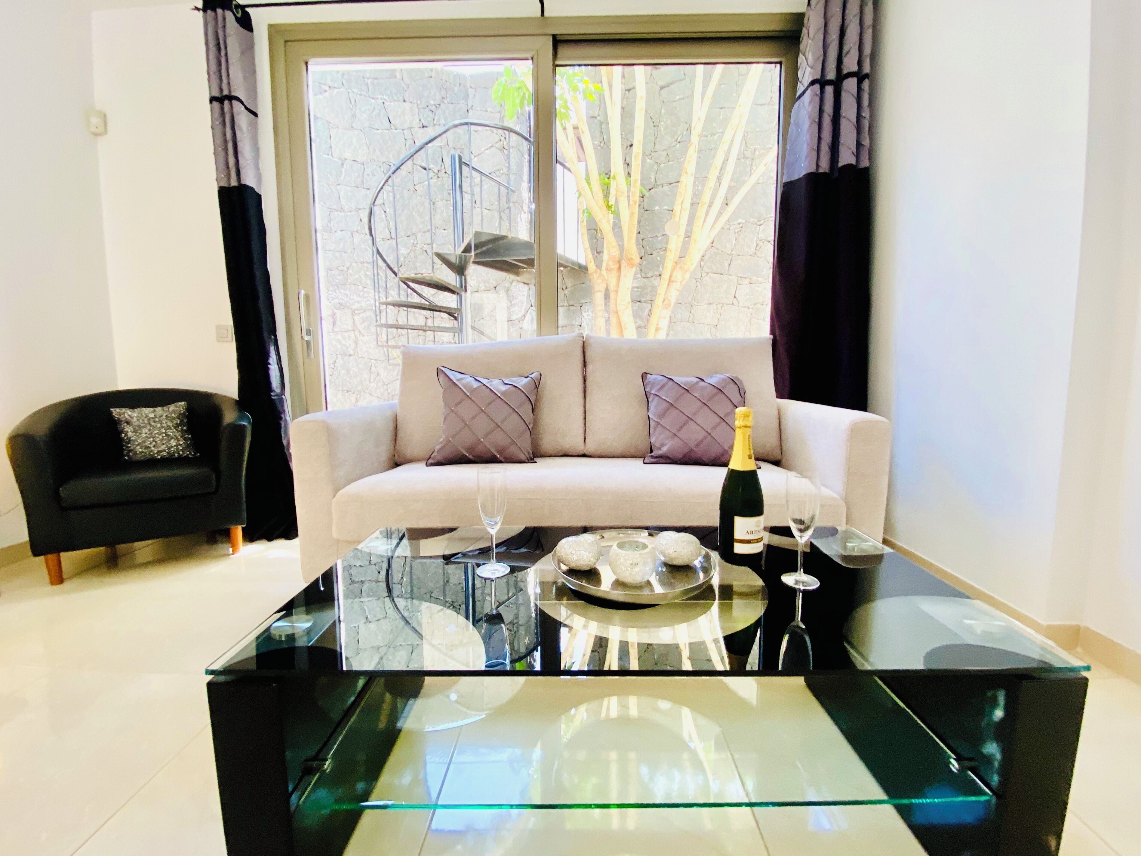 Ein modernes Wohnzimmer mit einem Glastisch, einem beige gefärbten Sofa mit gemusterten Kissen, einem schwarzen Sessel und einem großen Fenster mit Blick auf Bäume.