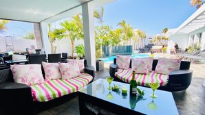 A covered outdoor seating area with wicker furniture, striped cushions, and a glass table with drinks and a bottle.