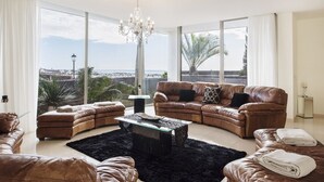 A spacious living room with leather sofas, a glass coffee table, and large windows offering a view of the sea.