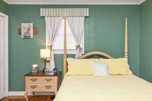 A bedroom with a four-poster bed, a nightstand with a lamp, and a window with curtains.