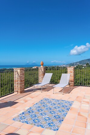Terraço com duas espreguiçadeiras, piso estampado e vista para o mar e montanhas ao longe.