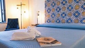 A neatly made bed with a folded towel and a book, a blue patterned wall, and a chair with a lamp in the background.