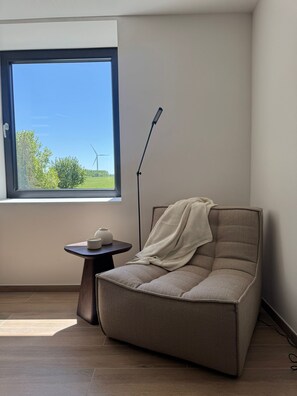 A modern room with a large window, a single chair, a small table, and a floor lamp. - Gite 3 in the heart of the countryside (Chimay)