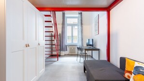 A room with a bed, a desk with a computer, a chair, a window with curtains, a radiator, and a staircase with red railings.