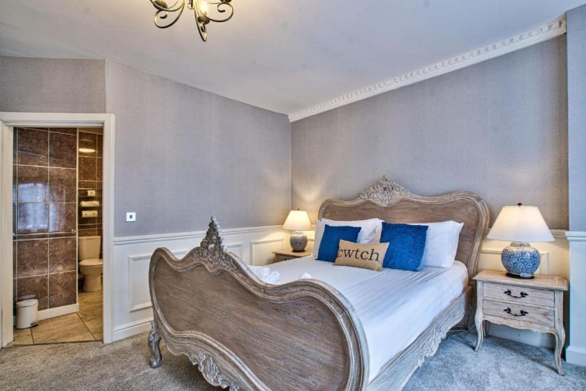 A bedroom with a large bed, two bedside tables with lamps, a chandelier, and a bathroom visible through an open door.
