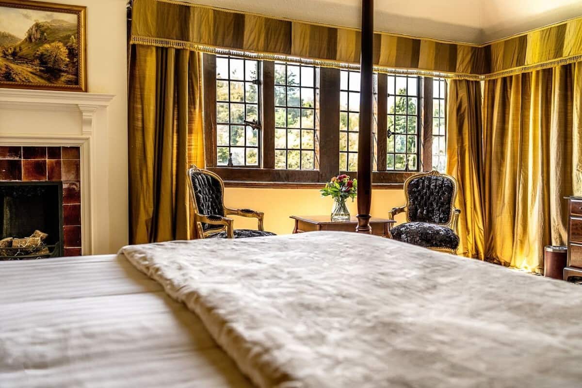 A room with a large bed, two chairs, a fireplace, and a window with curtains.