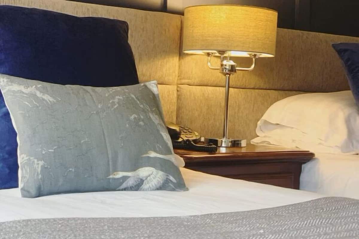 A neatly made bed with a grey textured bedspread, two pillows, a bedside lamp, and a wooden nightstand with a telephone.