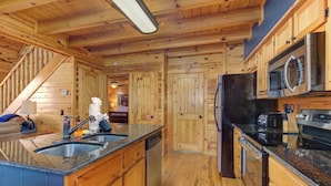 A wooden cabin interior with a kitchen area featuring a granite countertop, stainless steel appliances, and a wooden staircase.