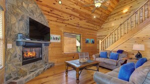 A cabin interior with a stone fireplace, wooden walls, a sofa, and a staircase.