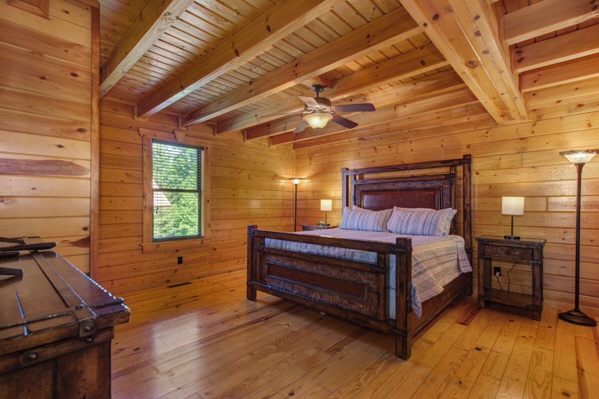 Een slaapkamer in een houten hut met een bed, een plafondventilator, een raam en een nachtkastje met een lamp.